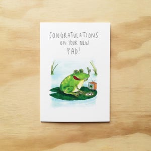May include: A white greeting card featuring a watercolour illustration of a green frog sitting on a lily pad. The frog is giving a thumbs up next to a small television set. The text reads: CONGRATULATIONS ON YOUR NEW PAD! in black ink.