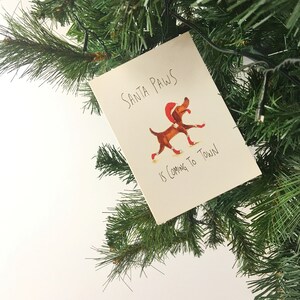 Puede incluir: Una tarjeta blanca con el texto "Santa Paws is coming to town" y una ilustraci&oacute;n de dibujos animados de un perro marr&oacute;n con un gorro de Pap&aacute; Noel rojo y botas rojas.