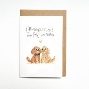 May include: A white greeting card with a brown envelope featuring a watercolour illustration of two brown dogs. One dog wears a black bow tie, the other a tiara and collar. Text reads: Congratulations You're PAWesome Together, with a small yellow heart.