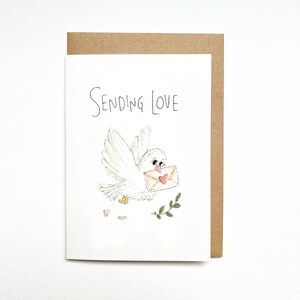 Può includere: Un biglietto d'auguri bianco con una busta marrone dietro. Il biglietto presenta un'illustrazione disegnata a mano di un uccello bianco in volo che tiene una busta color sabbia con un piccolo cuore rosso. Il testo "SENDING LOVE" è in nero.