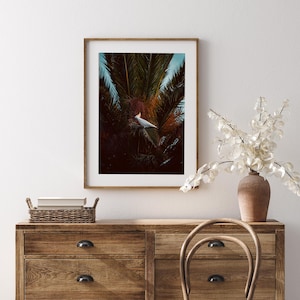May include: A framed wall art print featuring a white cockatoo with a yellow crest perched within dark green palm fronds. The artwork is displayed in a light wood frame hanging on a wall above a wooden dresser with a woven basket and a vase.