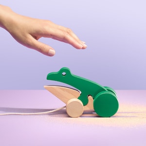 Könnte beinhalten: Ein grünes, hölzernes Spielzeug-Krokodil mit einer Zugschnur. Das Krokodil hat einen großen Kopf und einen langen Schwanz. Das Spielzeug befindet sich auf einer hellvioletten Oberfläche mit etwas Sand, der umher verstreut ist.