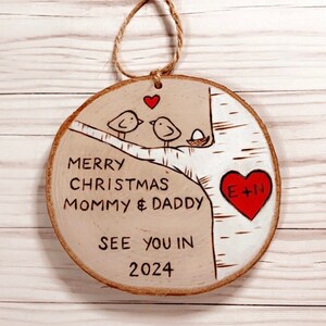May include: A wooden Christmas ornament with a birch tree design. Two birds are perched on a branch with a nest and egg. The ornament reads "Merry Christmas Mommy & Daddy See You in 2024" and has a red heart with "E+N" inside.