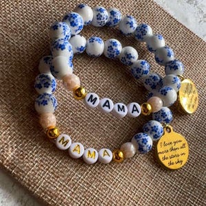 May include: Two beaded bracelets. One bracelet spells "MAMA" with white letter beads and gold accents. The other bracelet has white beads with blue floral patterns. Both bracelets have gold charms with text.