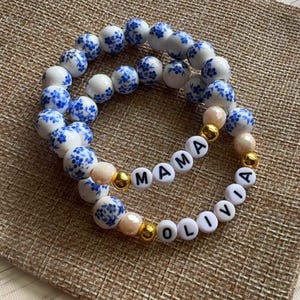 May include: Two beaded bracelets. One bracelet spells "MAMA" with white letter beads and gold accents. The other bracelet spells "OLIVIA". Both bracelets have white and blue floral beads and neutral-toned beads.