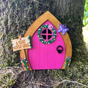 May include: A wooden fairy door with a pink door, a brown frame, and a green wreath. A sign reads "A Fairy Lives Here." A purple butterfly and pink flowers decorate the door. The door is mounted on a tree trunk.