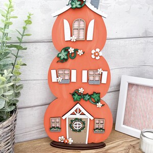 May include: A three-tiered pumpkin house decoration made of wood. The house is painted orange and has white windows and a brown door with a wreath. The house is decorated with green vines, white flowers, and a grey chimney.