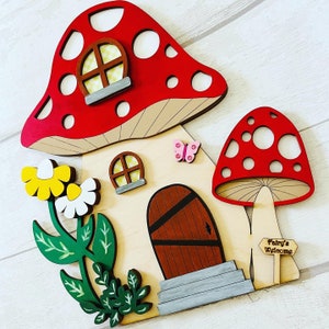 May include: A wooden fairy house with a red mushroom roof, a brown door, and a sign that says "Fairy's Welcome". The house has a flower with yellow and white petals and green leaves.