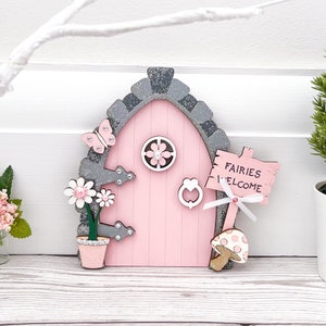May include: A pink fairy door with a grey stone frame, a sign that reads "Fairies Welcome", and a pink flower pot with a green plant.