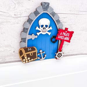 May include: A blue wooden fairy door with a skull and crossbones design, a treasure chest with gold coins, an anchor, and a ship's wheel. A red sign reads "A pirate lives here".