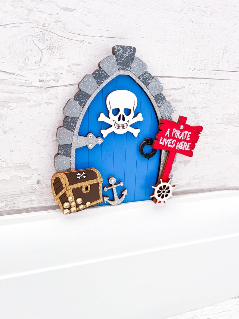 May include: A blue wooden fairy door with a skull and crossbones design, a treasure chest with gold coins, an anchor, and a ship's wheel. A red sign reads "A pirate lives here".