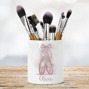 May include: White ceramic makeup brush holder with a pink watercolor design of ballet shoes and a bow. The holder has the name "Olivia" printed on the front.
