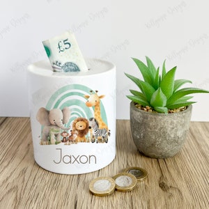 May include: White ceramic money box with a green and blue rainbow design. The box features a group of jungle animals including an elephant, lion, giraffe, zebra, monkey, and frog. The name "Jaxon" is printed on the front of the box.