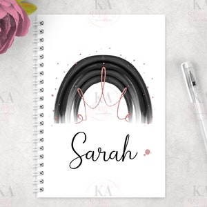 May include: A black and white rainbow with a pink crown and stars on a white background. The name "Sarah" is written in black cursive below the rainbow. The image is on a spiral bound notebook.