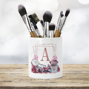 May include: White ceramic makeup brush holder with a rose gold geometric design and the letter "A" in the centre. The holder is filled with makeup brushes and has a pink floral design around the letter.
