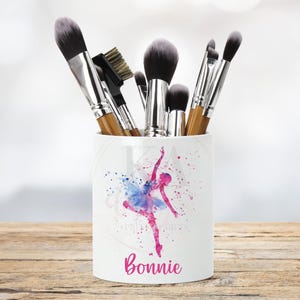 May include: White ceramic makeup brush holder with a watercolor design of a ballerina in a pink and blue tutu. The holder has the name "Bonnie" printed on the bottom.