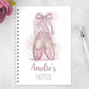 May include: A pink spiral bound notebook with a watercolour illustration of ballet shoes and a bow. The notebook says "Amelie's Notes" on the front.