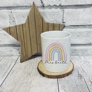 May include: White ceramic mug featuring a pastel rainbow design with pink, yellow, and blue arches. The mug has the name "Mrs Booth" written in a script font. The mug sits on a wooden coaster, with a wooden star in the background.