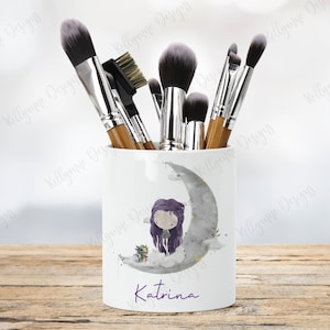 May include: White ceramic makeup brush holder with a whimsical illustration of a girl sitting on a crescent moon with a small dragon. The holder has the name "Katrina" written in purple cursive at the bottom.