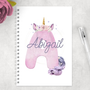 May include: A pink unicorn notebook with a floral crown and the name Abigail written in purple. The notebook has a spiral binding and is perfect for taking notes or journaling.