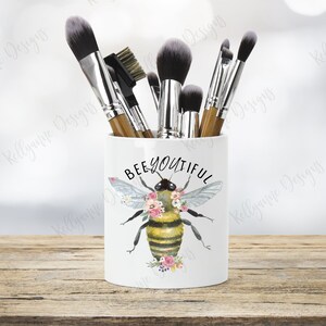 Peut inclure: Porte-pinceaux de maquillage en céramique blanche avec un motif d'abeille et de fleurs. Le texte "BEE-YOU-TIFUL" est imprimé sur le support. Le support est rempli de pinceaux de maquillage.