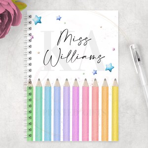 May include: A spiral-bound notebook with the name "Miss Williams" in elegant script. The cover features colorful pencils at the bottom, star accents, and a pen resting beside it. A pink flower is in the upper left corner.