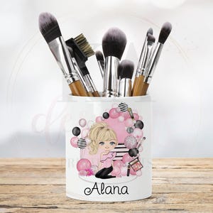 May include: White ceramic makeup brush holder with a pink and black design featuring a cartoon girl with the name "Alana" printed on the front.