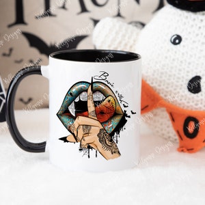 May include: White ceramic mug with a black interior and handle. The mug features a colorful design of lips with a vampire fang, a hand with a finger to the lips, and the text "Basic witched". The design includes bats and spiderwebs.