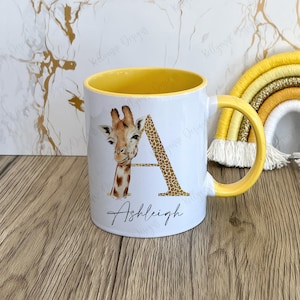 Peut inclure: Une tasse en céramique blanche avec un bord et une poignée jaunes. La tasse présente une lettre "A" dorée avec une girafe qui se cache derrière. Le nom "Ashleigh" est écrit sous la lettre.
