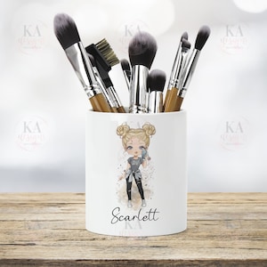 May include: White ceramic makeup brush pot featuring a cartoon girl illustration and the name "Scarlett". The pot holds several makeup brushes with wooden handles and black bristles. The background is blurred.