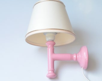Op de afbeelding: Een roze keramische wandlamp met een witte lampenkap van stof. De lampenkap heeft een gouden rand rond de bovenrand. De wandlamp heeft een eenvoudig ontwerp met een T-vormige arm en een ronde basis.