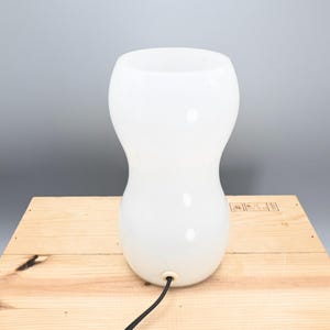 May include: A white ceramic table lamp with a rounded, hourglass-shaped base. The lamp has a black cord.