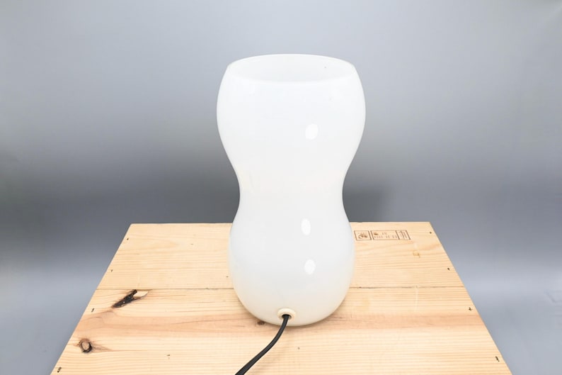 May include: A white ceramic table lamp with a rounded, hourglass-shaped base. The lamp has a black cord.