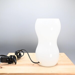 May include: A white glass table lamp with a rounded, hourglass-shaped base. The lamp has a black cord and a switch.