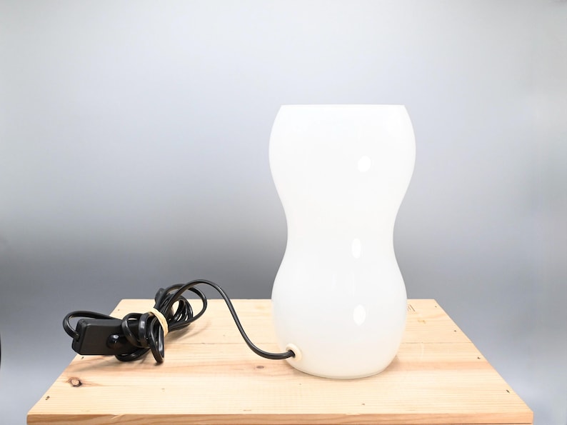May include: A white glass table lamp with a rounded, hourglass-shaped base. The lamp has a black cord and a switch.