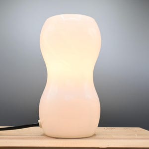 May include: A white ceramic table lamp with a rounded, hourglass-shaped base. The lamp is lit and casting a warm glow.