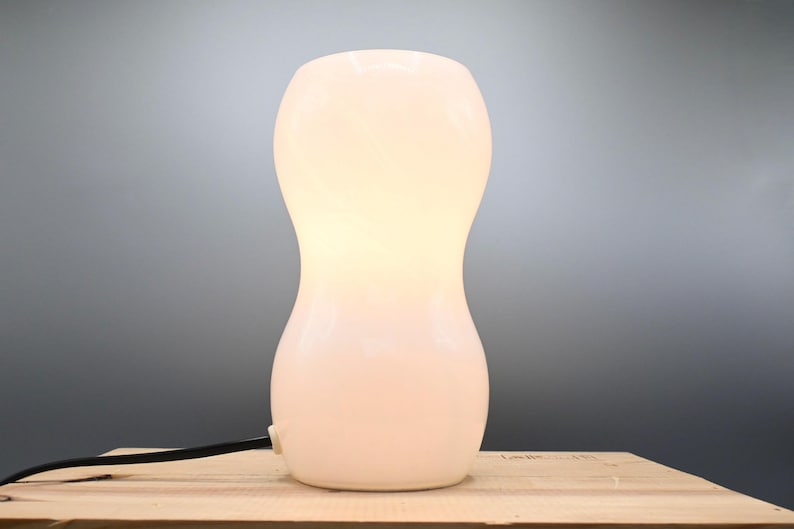 May include: A white ceramic table lamp with a rounded, hourglass-shaped base. The lamp is lit and casting a warm glow.