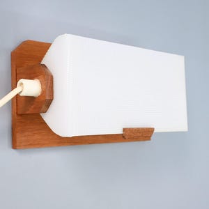 Può includere: Una lampada da parete con base rettangolare in legno e paralume in vetro bianco testurizzato con motivo a griglia. La lampada include un cavo elettrico di colore chiaro che fuoriesce dal lato, collegato a un attacco bianco.