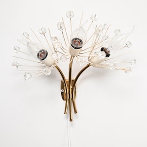 Op de afbeelding: Een gouden en witte wandlamp met drie lampen en kristallen accenten. De lamp is gevormd als een gestileerde bloem met drie takken.