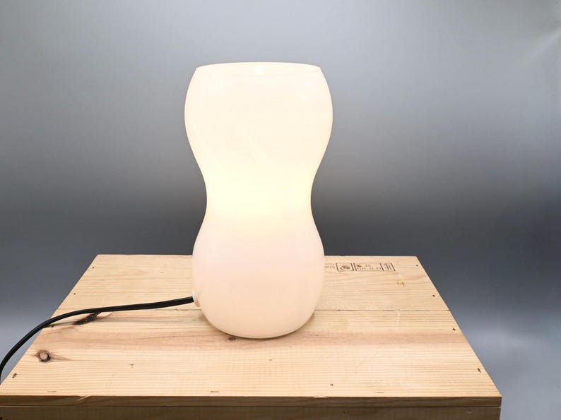 May include: A white, hourglass-shaped table lamp with a smooth, frosted glass shade. The lamp is turned on and illuminating the room.