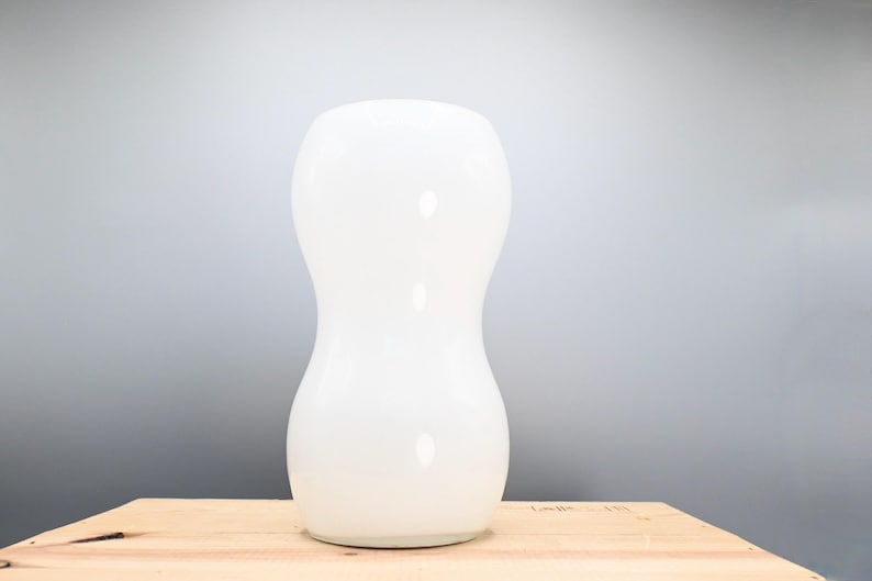 May include: A white ceramic vase with a rounded, hourglass shape. The vase is sitting on a wooden surface.