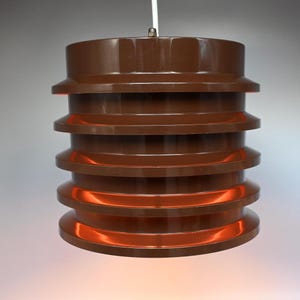 Może przedstawiać: Brązowa, wielowarstwowa lampa wisząca z ciepłym pomarańczowym światłem. Lampa jest wykonana z tworzywa sztucznego i ma nowoczesny design.
