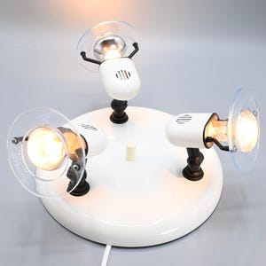 Puede incluir: Una luminaria de riel blanca de tres luces con pantallas de vidrio transparente y detalles negros. La luminaria tiene una base redonda y un cable para enchufar.