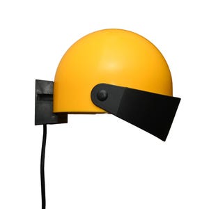 Peut inclure: Une lampe murale jaune semi-sphérique avec une visière et un support de montage noirs. La lampe a un cordon d'alimentation noir et est placée sur un fond blanc. Le design est moderne et minimaliste.