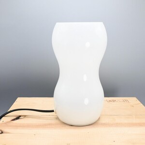 May include: White ceramic table lamp with a rounded, hourglass-shaped base. The lamp has a black cord.