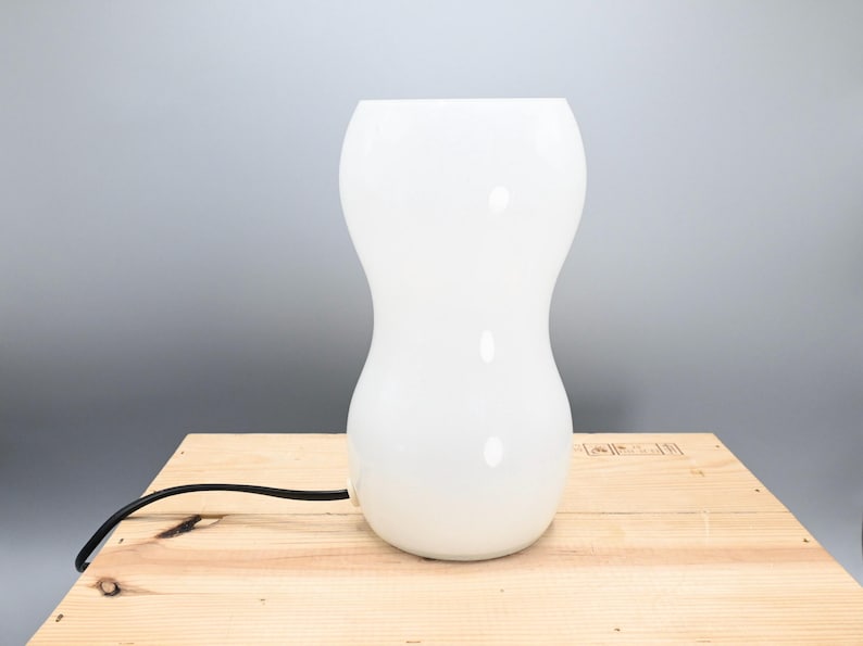May include: White ceramic table lamp with a rounded, hourglass-shaped base. The lamp has a black cord.
