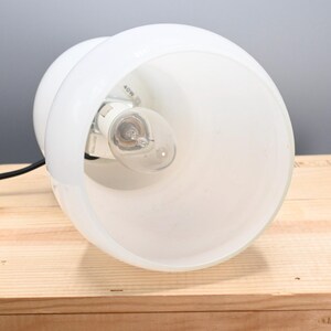 May include: White table lamp with a frosted glass shade and a black cord. The lamp is on a wooden surface.
