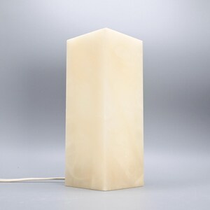 May include: A rectangular, cream-colored lamp with a square base and top. The lamp has a soft, diffused glow and a power cord. The lamp is a decorative lighting fixture.