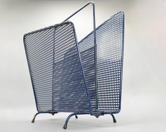 Puede incluir: Un revistero de metal azul con un patrón de rejilla. El revistero tiene un diseño único en ángulo y cuatro patas.