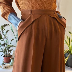 Könnte beinhalten: Nahaufnahme einer braunen Hose mit hohem Bund und einzigartigen Taschendetails. Die Hose hat ein weites Bein und eine maßgeschneiderte Passform. Das Model trägt einen braunen Pullover mit blauen und weißen Akzenten an den Ärmeln. Die Hose ist aus einem glatten, einfarbigen Stoff gefertigt.